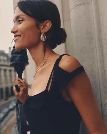 Portrait of a woman wearing a black dress and gold jewelry smiling on a balcony with an urban cityscape in the background.
