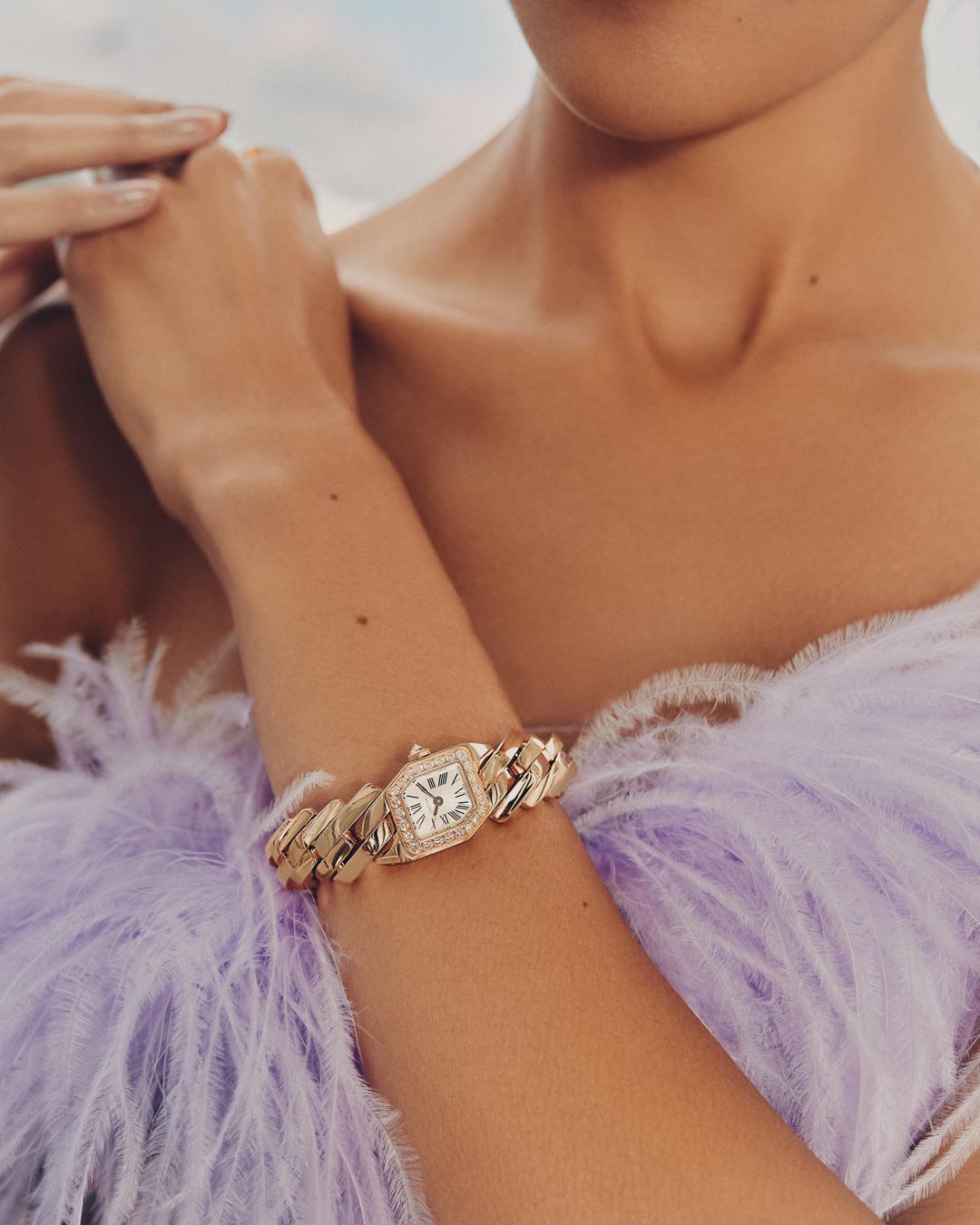 Close-up of a woman's arm with a gold chain-link watch, adorned with diamonds, against a pale blue and white background with soft lilac feathers.
