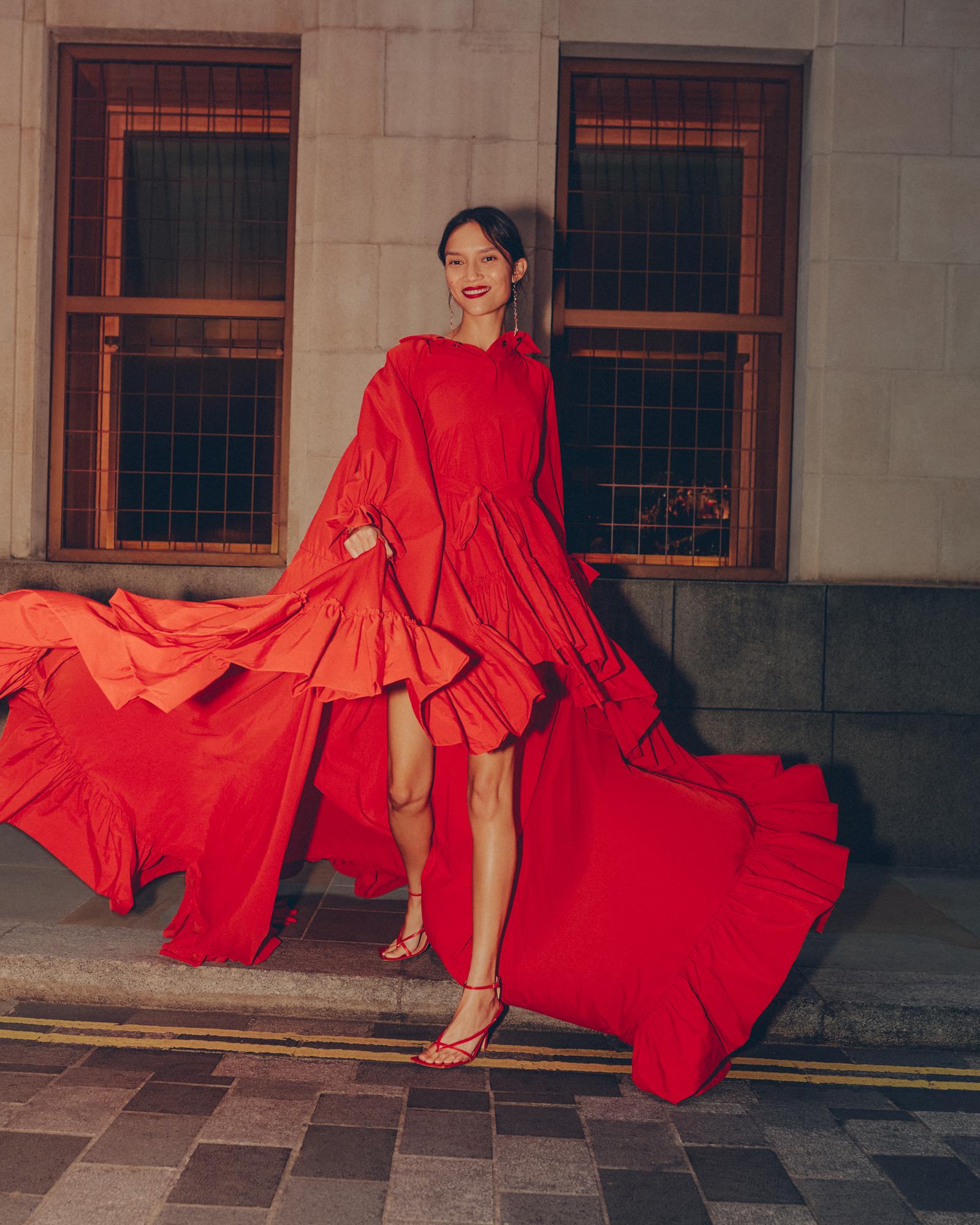 A woman in a vibrant red, layered dress, posing outdoors against a neutral building with window details, with a striking, fashionable appearance.