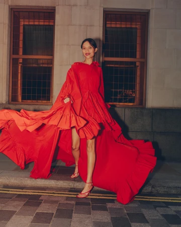 A woman in a vibrant red, layered dress, posing outdoors against a neutral building with window details, with a striking, fashionable appearance.