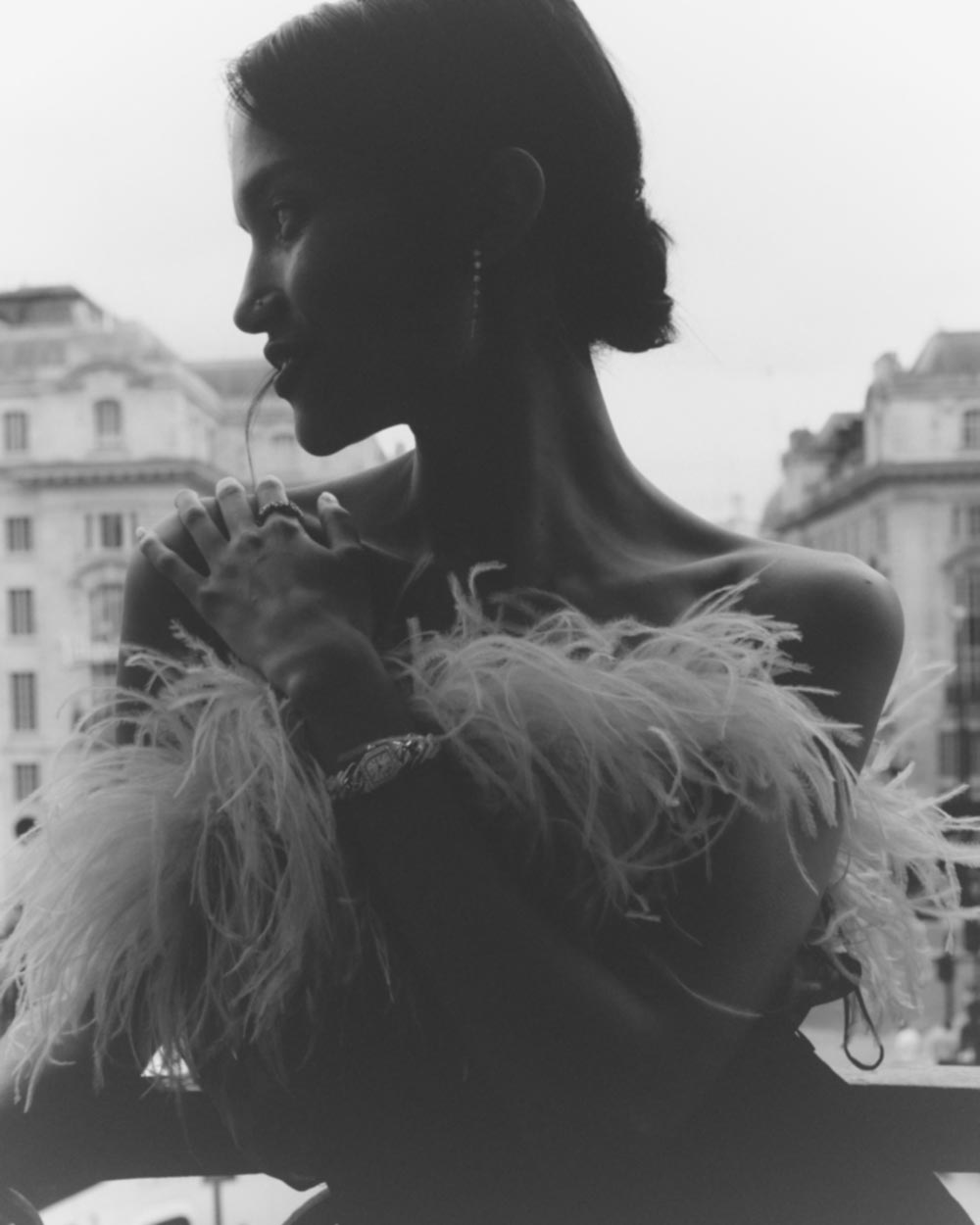 Monochrome close-up of a woman in a feathered garment with blurred city buildings and cloudy skies in the background.