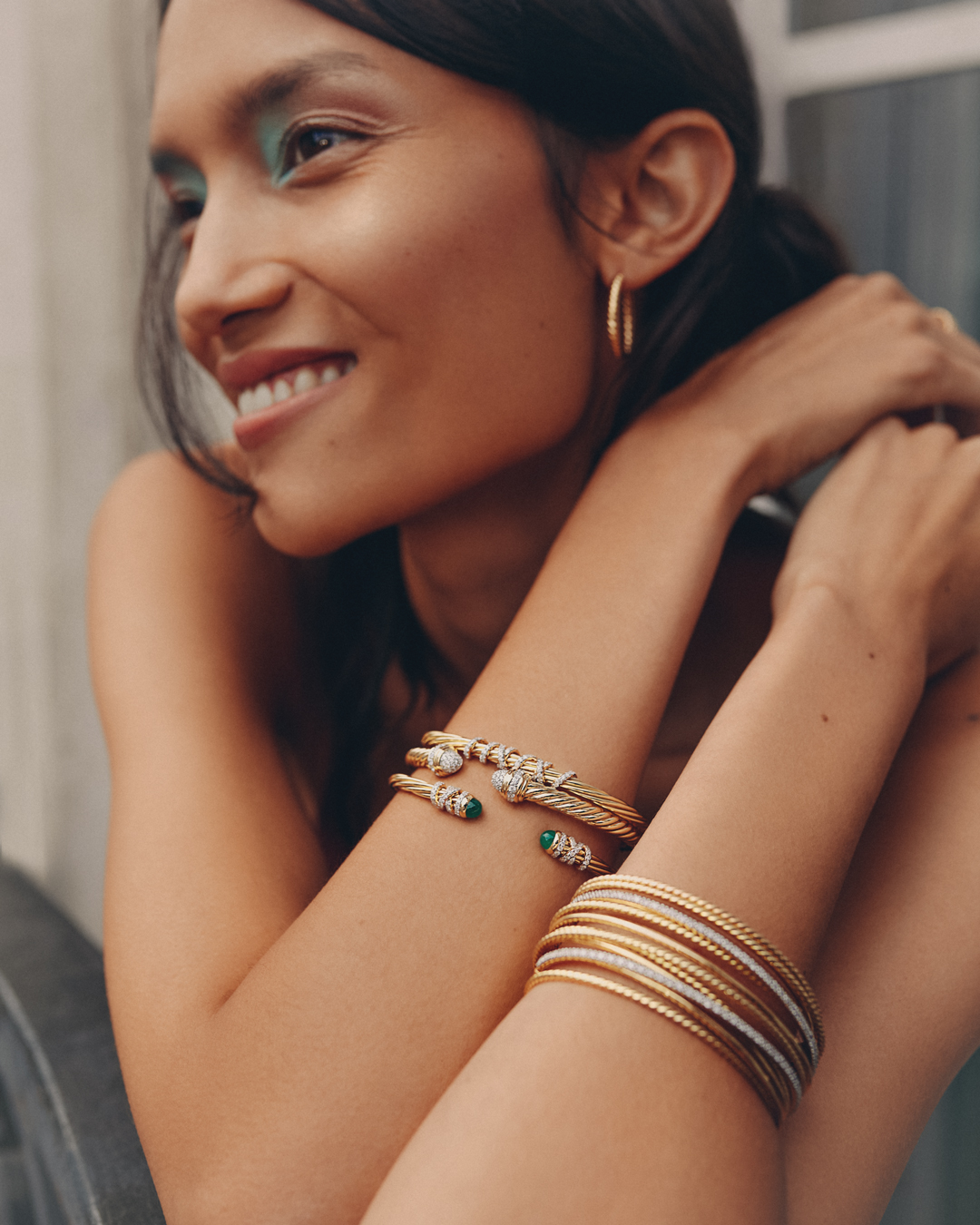 Adult female smiles with gold hoop earrings and bracelets on her arms in a studio setting with filtered natural light.