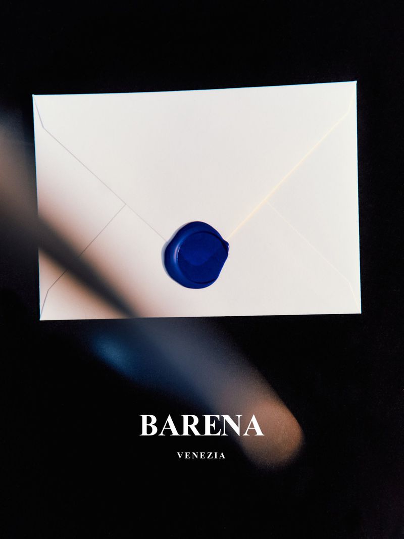 Angled view of a white envelope with a blue wax seal on a dark background, with a light beam and the Barena Venezia logo.