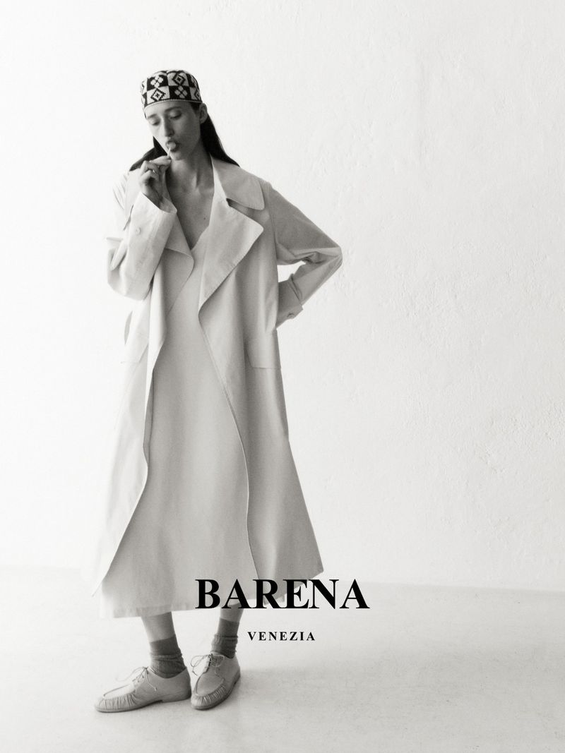 Black and white portrait of a woman wearing a patterned cap and long coat, standing indoors against a bright white background.