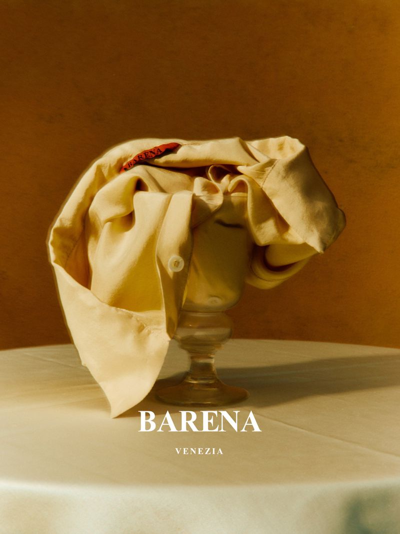 Still life shot of a folded cream-colored shirt on top of a stemmed glass against a warm brown background with brand logo visible at the bottom.