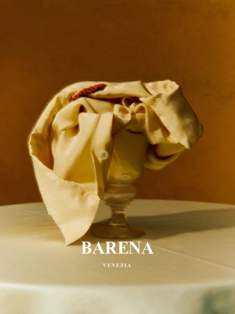 Still life shot of a folded cream-colored shirt on top of a stemmed glass against a warm brown background with brand logo visible at the bottom.