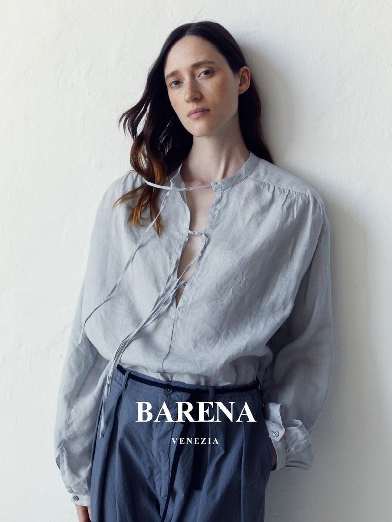 Elegant woman in a casual grey long-sleeve shirt and blue trousers posing against a white wall in a studio setting, fashion advertisement.