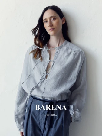 Elegant woman in a casual grey long-sleeve shirt and blue trousers posing against a white wall in a studio setting, fashion advertisement.