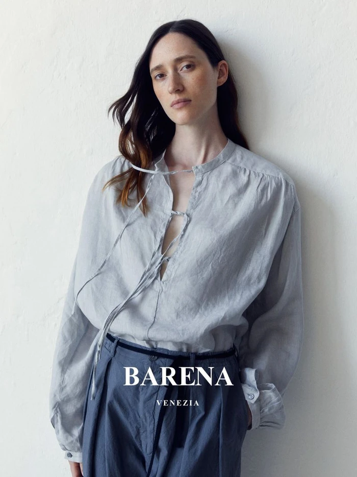 Model wearing a light grey tie-front blouse and blue trousers leaning against a white textured wall