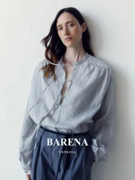 Model wearing a light grey tie-front blouse and blue trousers leaning against a white textured wall