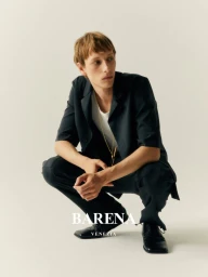 A male model crouching in a dark suit jacket, trousers, and black shoes against a plain white studio background.