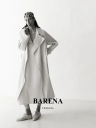 Model wearing a long light-colored coat and patterned headscarf against a plain white studio background.