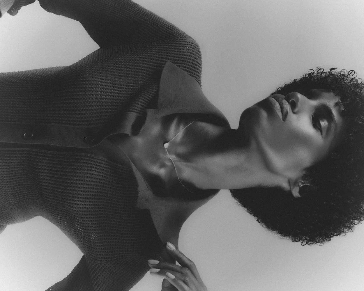 Monochrome portrait of an adult female with curly hair wearing a button-down shirt, posing against a light background in a studio.