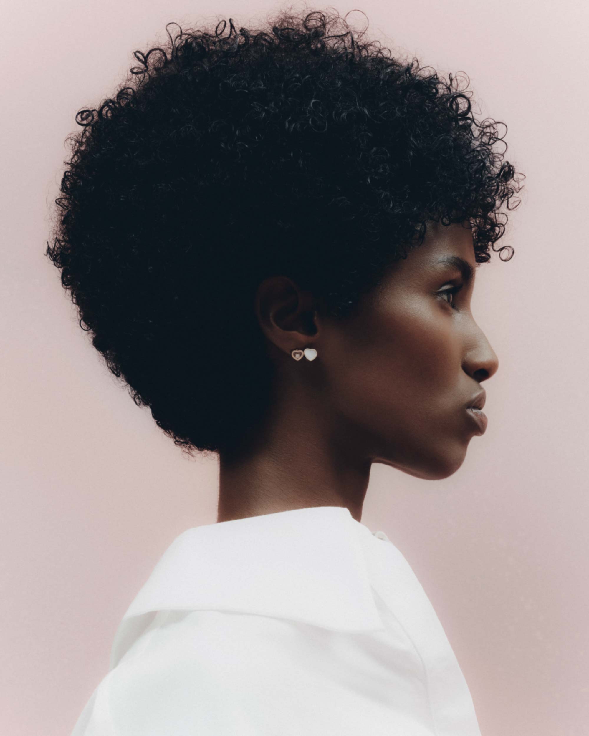 Profile portrait of an African American woman with curly afro hair, wearing a white collared shirt, against a soft pink background.