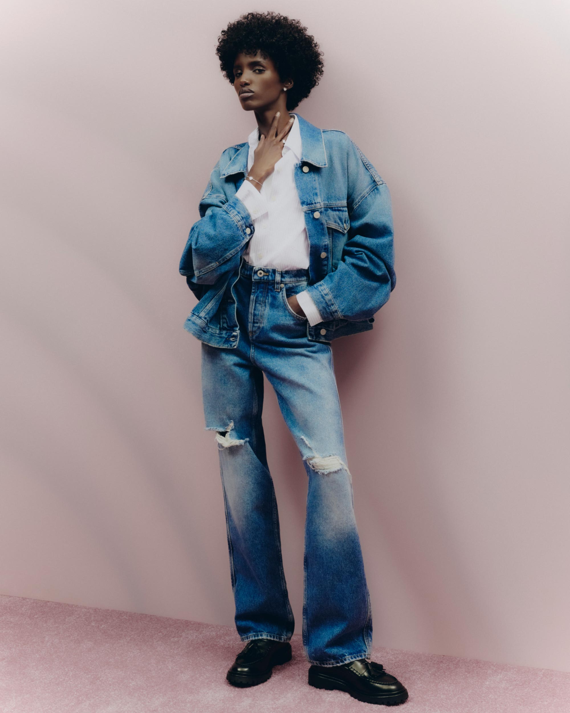 Fashion portrait of an African American female model wearing a denim jacket, jeans, white shirt, and black loafers against a pink background.