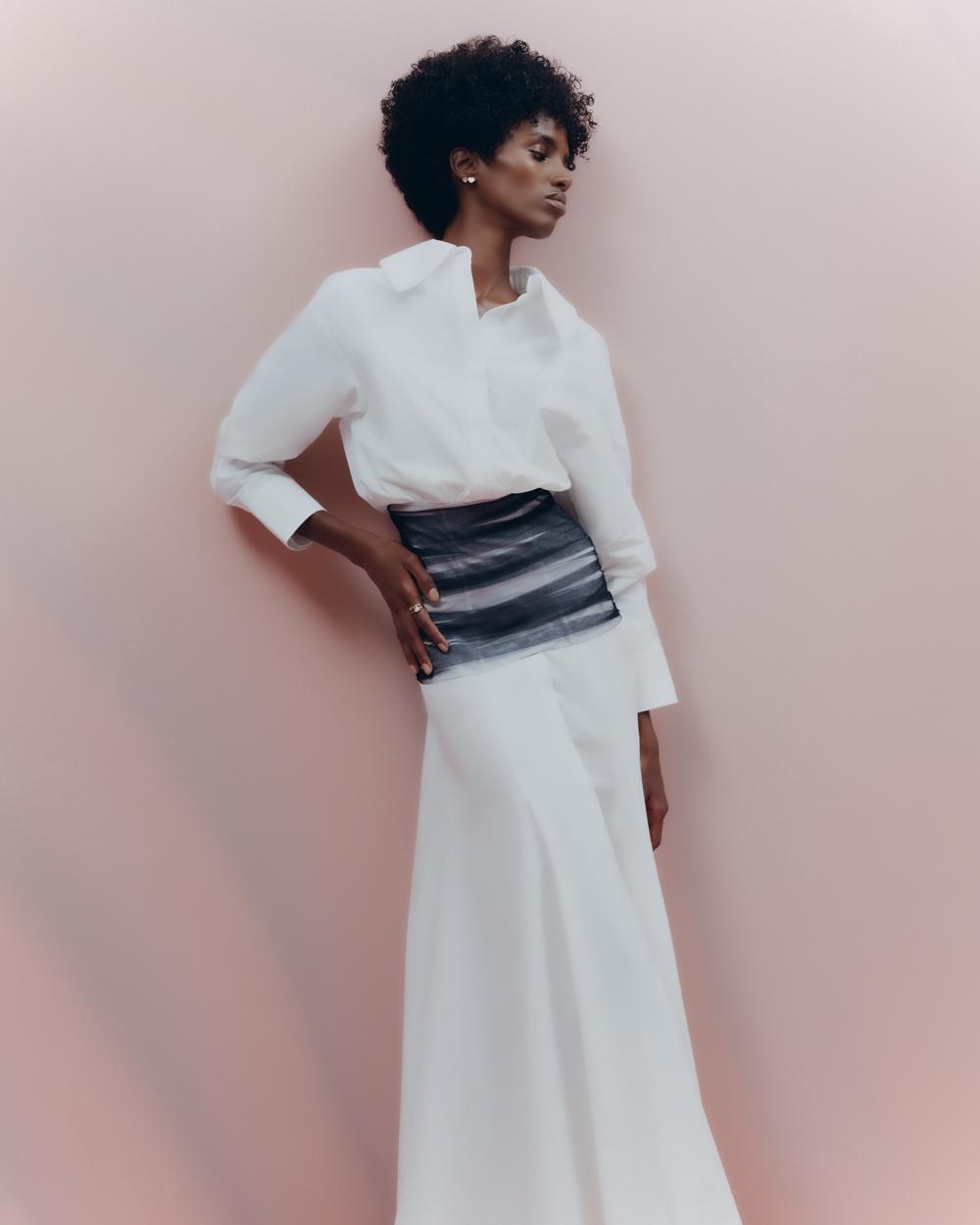 Elegant Black woman in flowing white skirt and blouse poses against a light pink background, showcasing a stylish fashion ensemble.