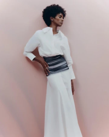 Elegant Black woman in flowing white skirt and blouse poses against a light pink background, showcasing a stylish fashion ensemble.