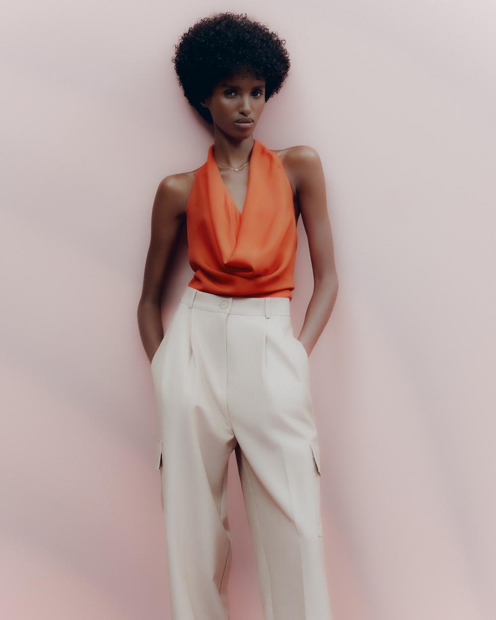 Fashion model in a bright orange cowl neck top and light beige high-waisted trousers, posing against a light pink background.