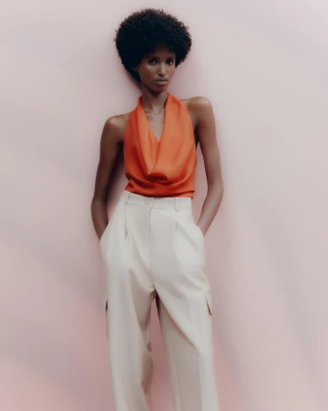 Fashion model in a bright orange cowl neck top and light beige high-waisted trousers, posing against a light pink background.