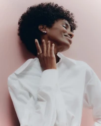 Close-up portrait of a Black woman with short dark curly hair wearing a white shirt and delicate jewelry against a soft pink backdrop.