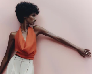 Model in an orange halter top and light trousers posing against a solid pink backdrop