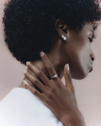 Close-up portrait of a Black woman wearing diamond earrings and a ring, with her hand resting on her neck.