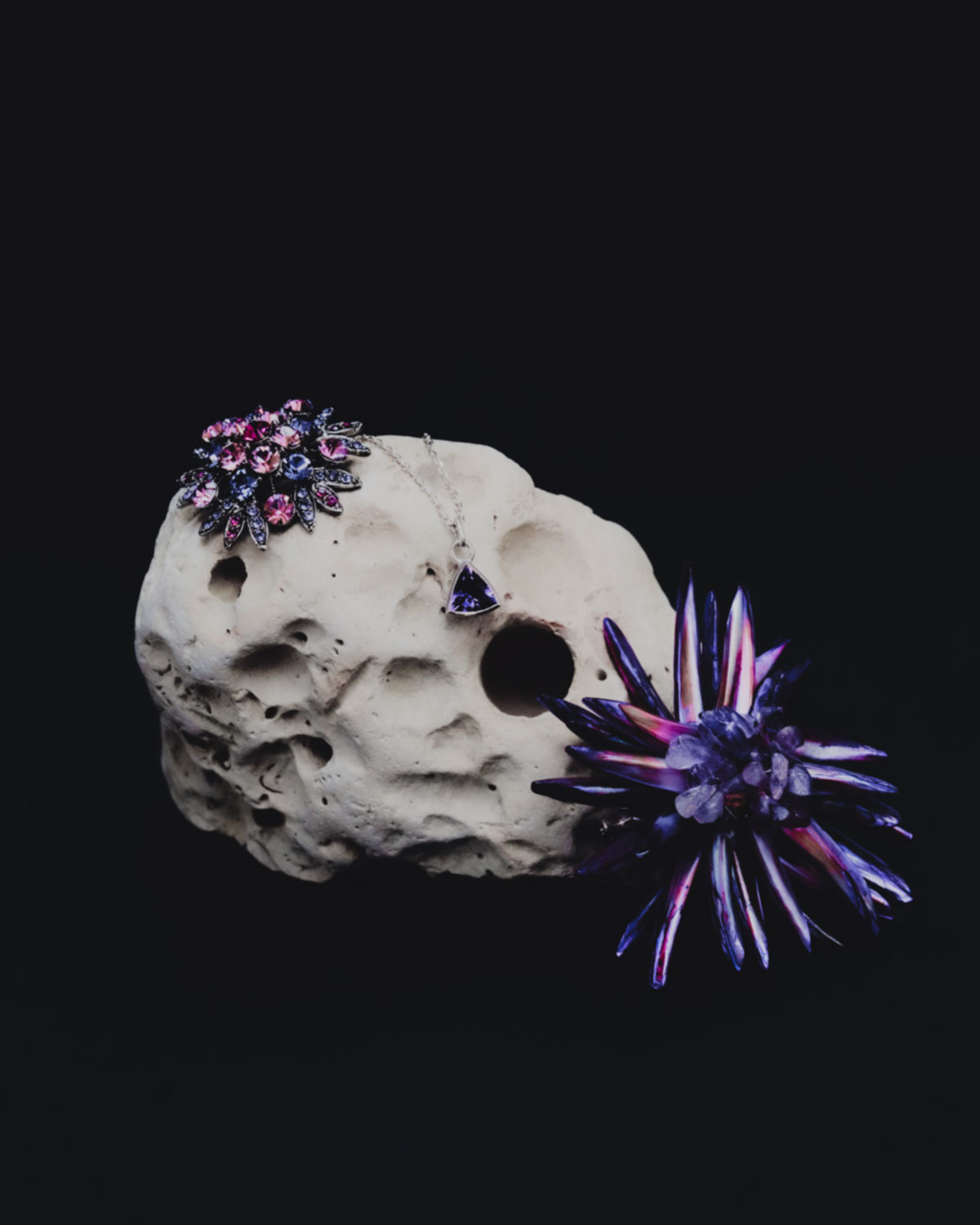 Jewellery and decorative flowers arranged on a textured stone platform against a dark background in a studio setting, creative product shot.
