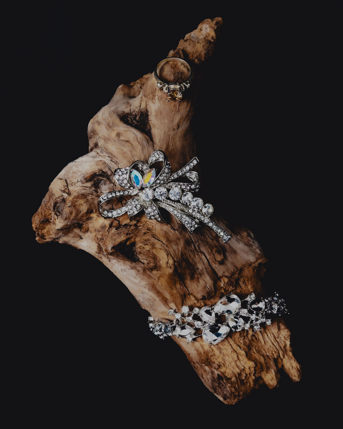 Silver and crystal jewelry pieces including a ring, brooch, and bracelet displayed on a textured driftwood branch against a dark backdrop.