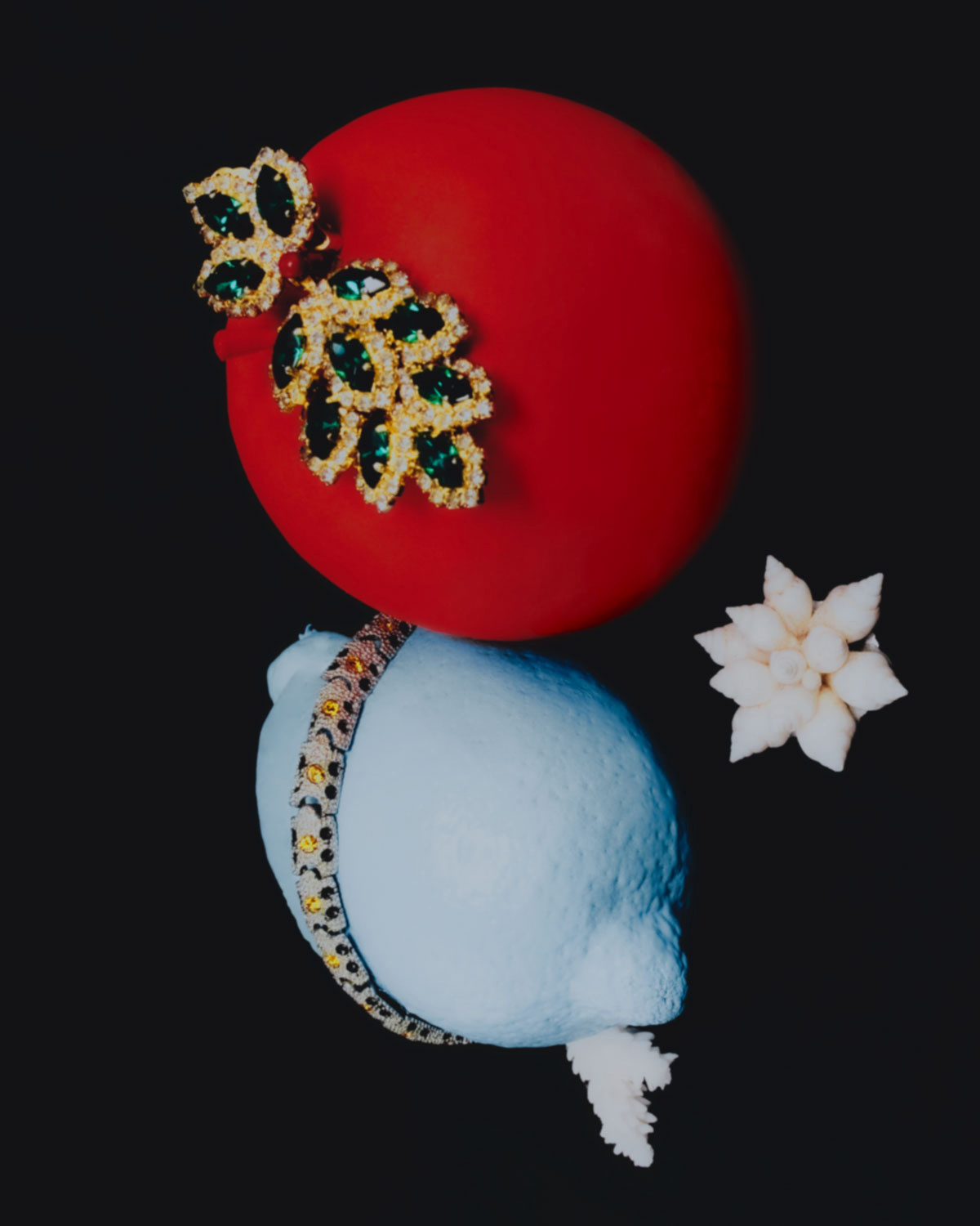 Still life of a red Christmas bauble with a decorative gold and green jewel, atop a light blue lemon, against a dark backdrop.