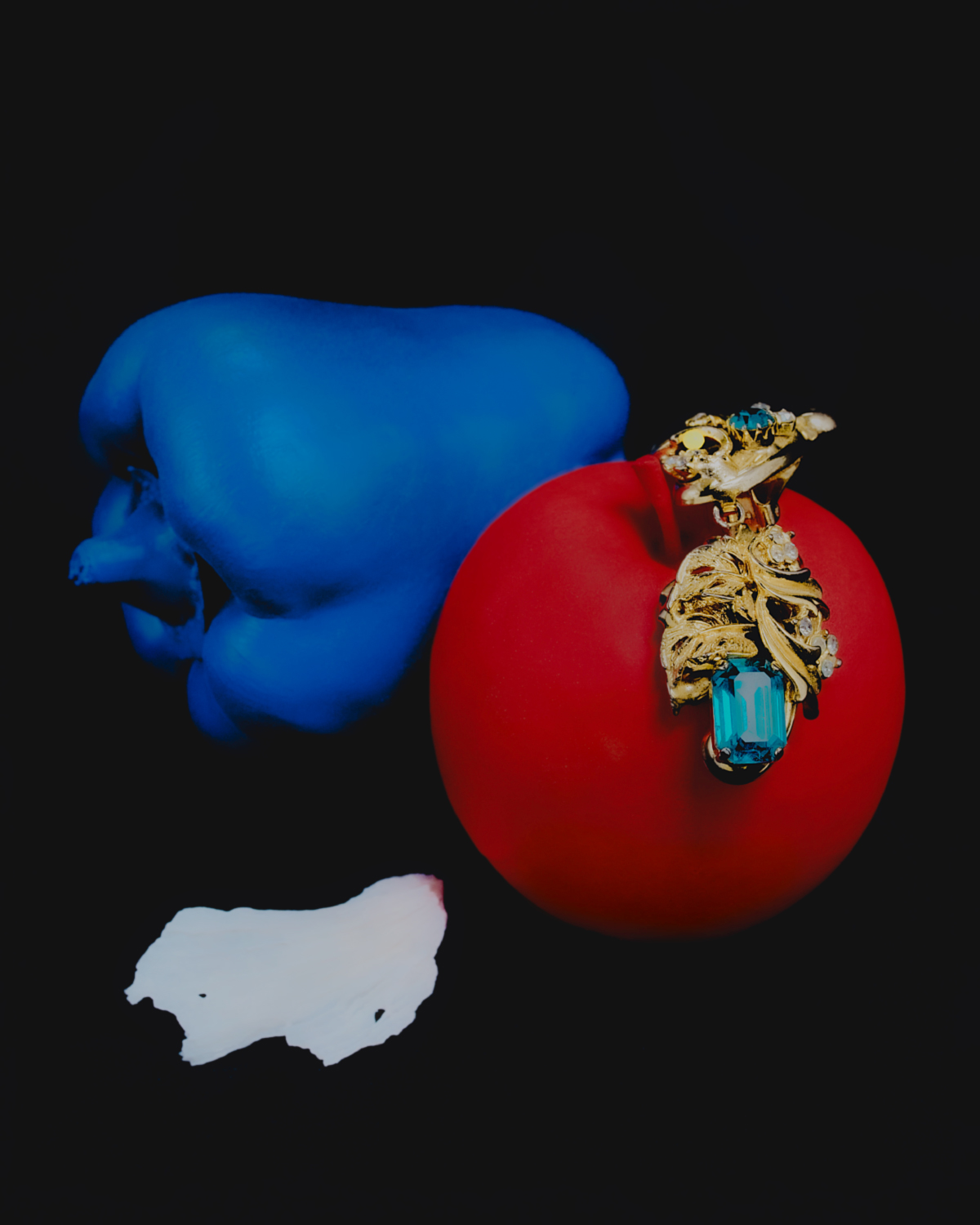 Still life photograph of red apple and blue pepper, with a gold pendant and a white paint splash on a black background.