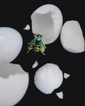 A vibrant insect-shaped brooch, sparkling in shades of green and gold, rests amidst the stark white fragments of a cracked egg.