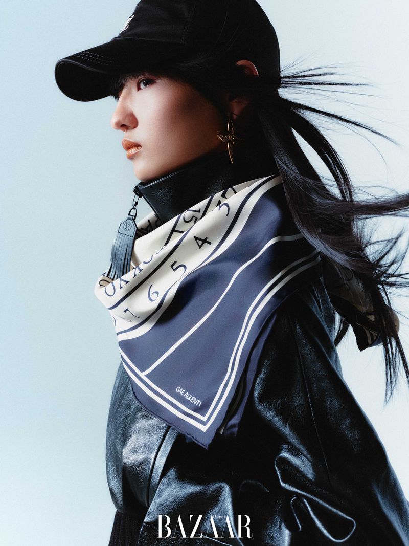 Asian woman wearing a black baseball cap, leather jacket, and patterned silk scarf poses in a fashion studio with a light blue background.