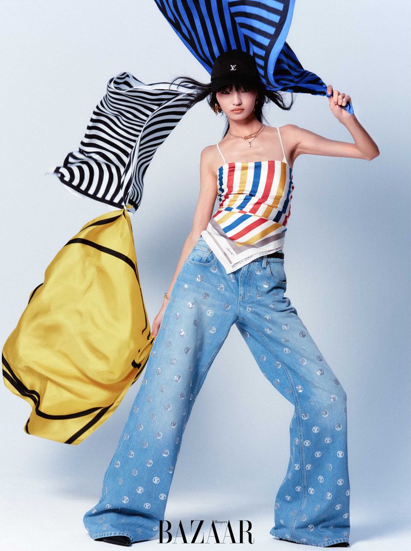 A fashionable young woman in a striped top and patterned jeans poses in a photo studio surrounded by flowing striped scarves.
