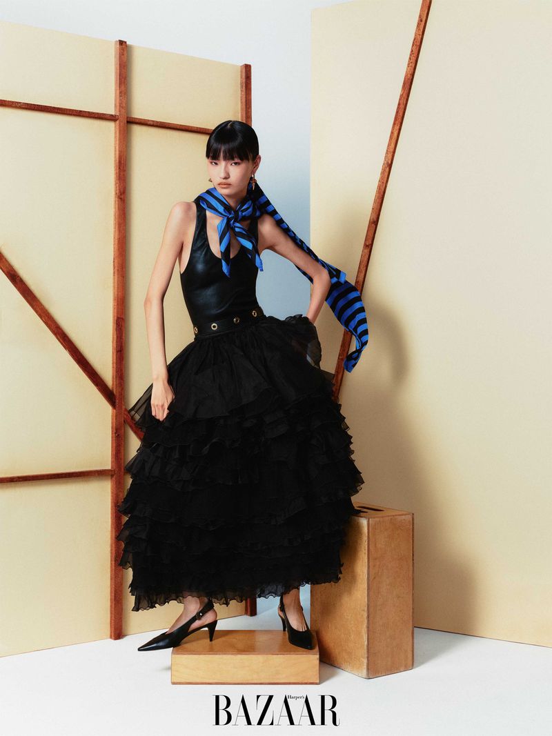 A stylish Asian woman poses indoors in a black top, tulle skirt, and heels, accessorized with a blue scarf, set against geometric backdrop.