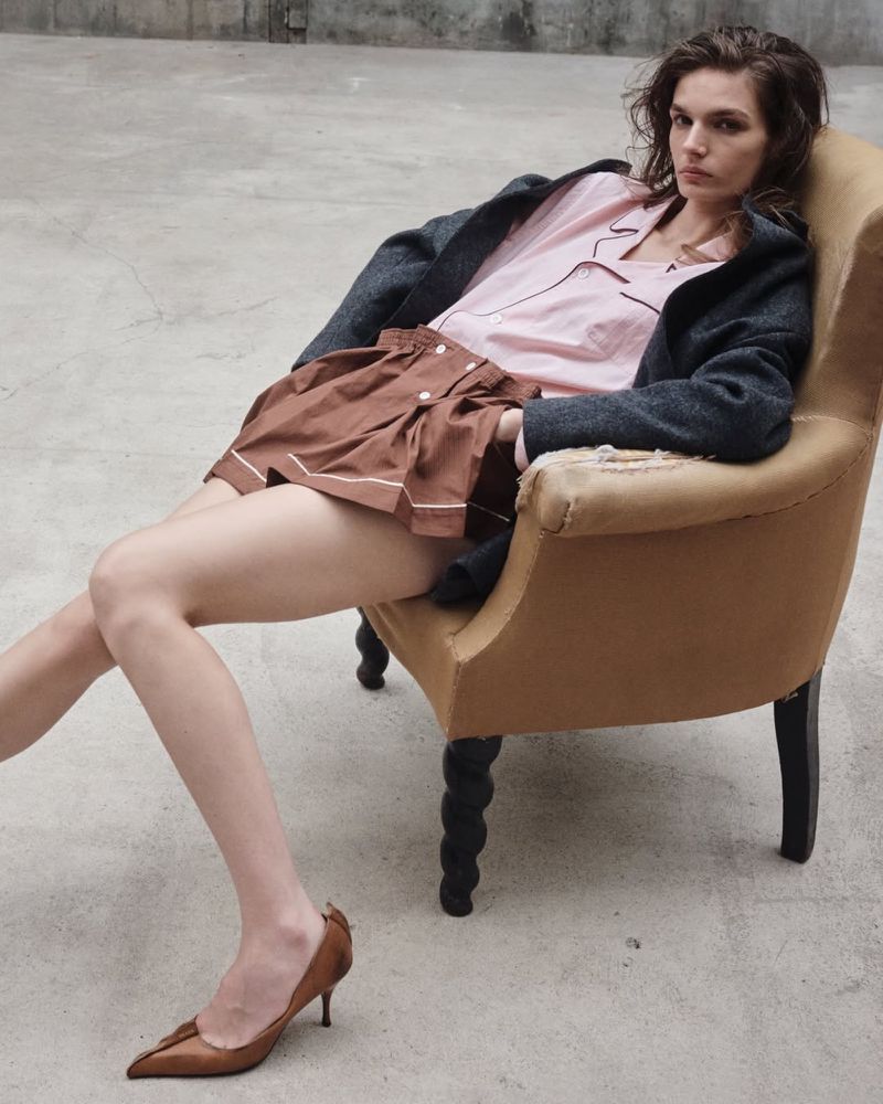 A model sits in a vintage chair wearing a pink pajama-style top, brown shorts, a grey jacket, and brown heels on a grey concrete floor.