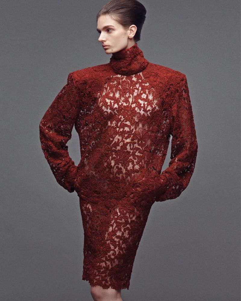 A slender fashion model poses in a semi-transparent, long-sleeved, dark-red, floral lace dress against a plain grey backdrop.
