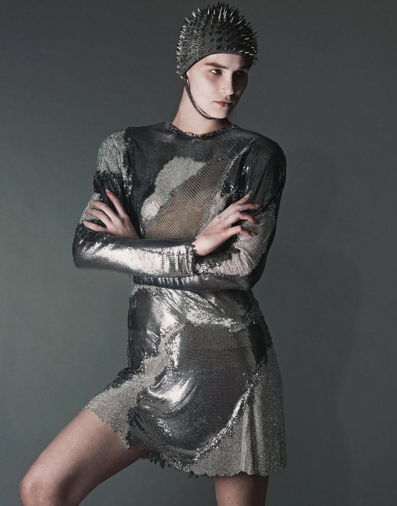 Adult female wearing metallic, long-sleeved mini dress and studded cap, arms crossed, against a grey backdrop in a studio fashion shot.