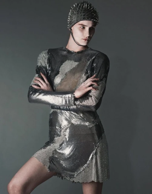 Adult female wearing metallic, long-sleeved mini dress and studded cap, arms crossed, against a grey backdrop in a studio fashion shot.