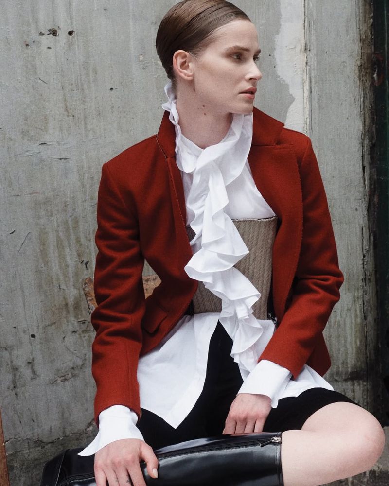 A fashionable adult female with dark hair poses sitting outdoors wearing a red jacket, white ruffled shirt, corset, and black boots.