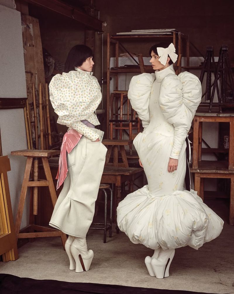 Two fashion models in puffy couture dresses and sculptural heels pose in an artist's studio surrounded by wooden stands and props.