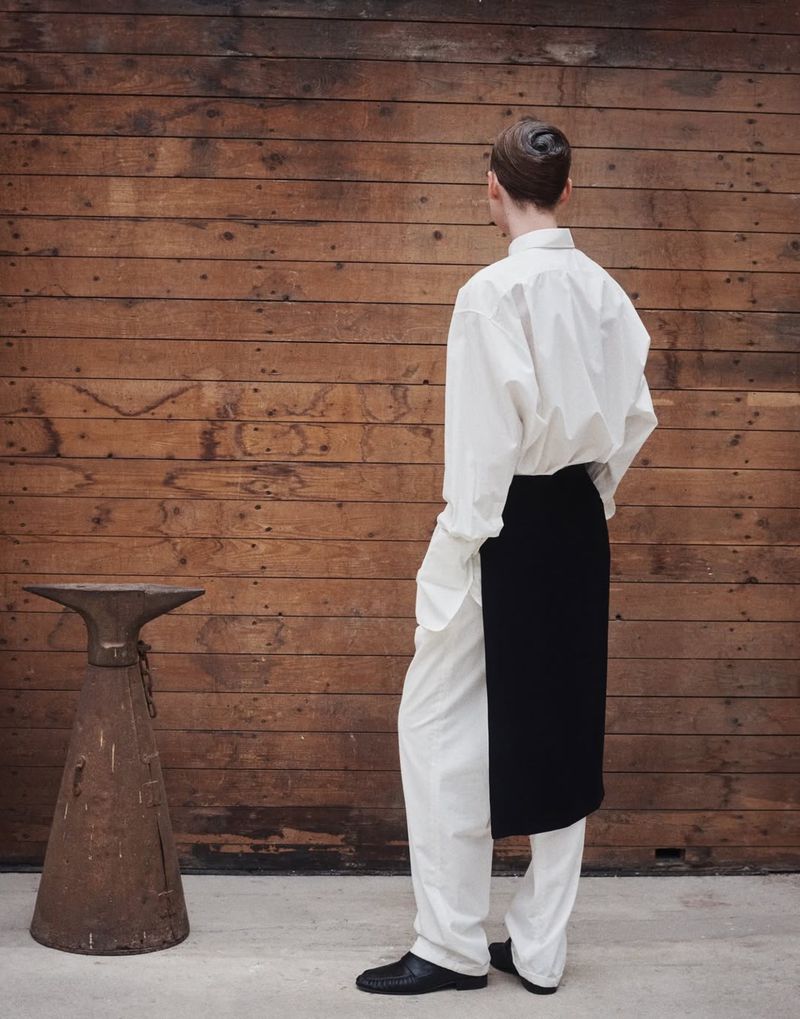 Back view of adult in white shirt and pants, black apron, and black shoes, standing near a rustic metal object and wood wall.