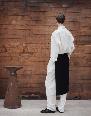 Back view of adult in white shirt and pants, black apron, and black shoes, standing near a rustic metal object and wood wall.