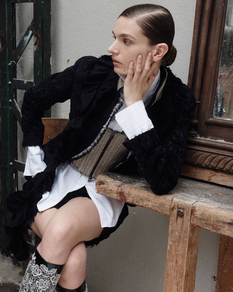 Woman in black coat, white shirt, and floral boots sits on a wooden structure. She's resting her face on her hand.