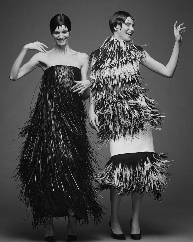Two smiling women pose in black and white gowns with fringe and feather details against a grey backdrop, capturing a fashion-forward style.