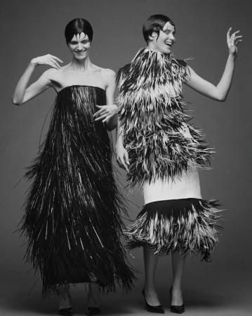 Two smiling women pose in black and white gowns with fringe and feather details against a grey backdrop, capturing a fashion-forward style.