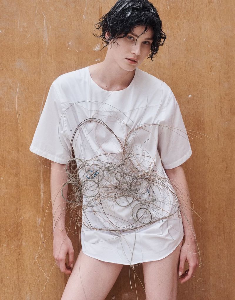 Artistic portrait of a young adult wearing a white t-shirt with a wire design, posing against a neutral brown wall in a photo studio.