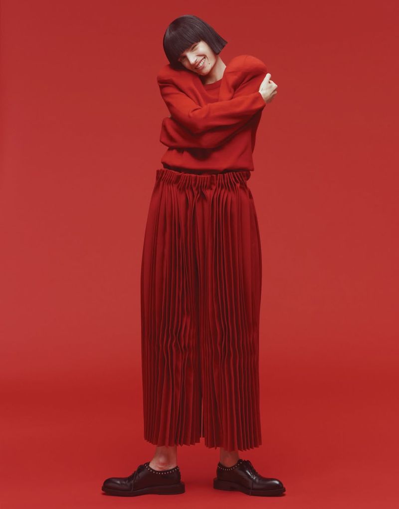 Woman in red sweater and pleated pants posing against a red background in a studio, looking at camera with soft, diffused lighting.