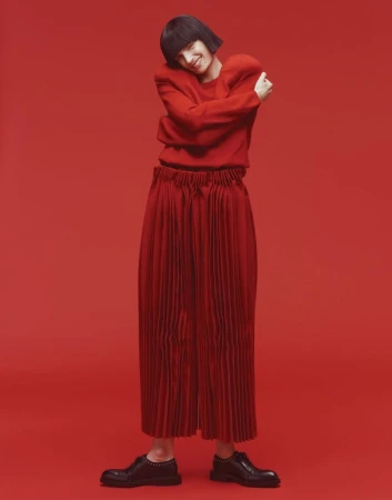 Woman in red sweater and pleated pants posing against a red background in a studio, looking at camera with soft, diffused lighting.