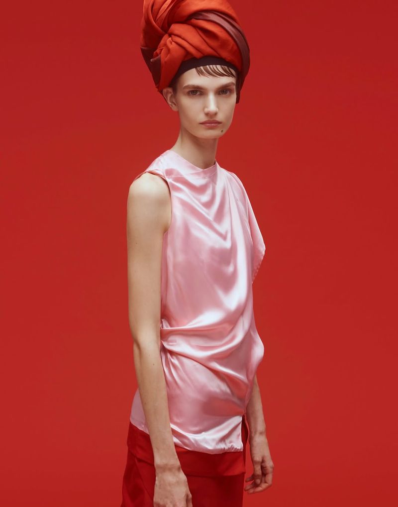 A fair-skinned woman with a red head wrap poses in a pink satin top and red pants against a red backdrop in a studio setting.