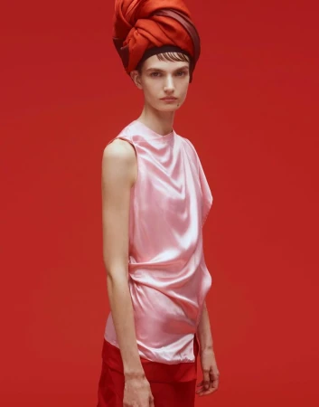 A fair-skinned woman with a red head wrap poses in a pink satin top and red pants against a red backdrop in a studio setting.